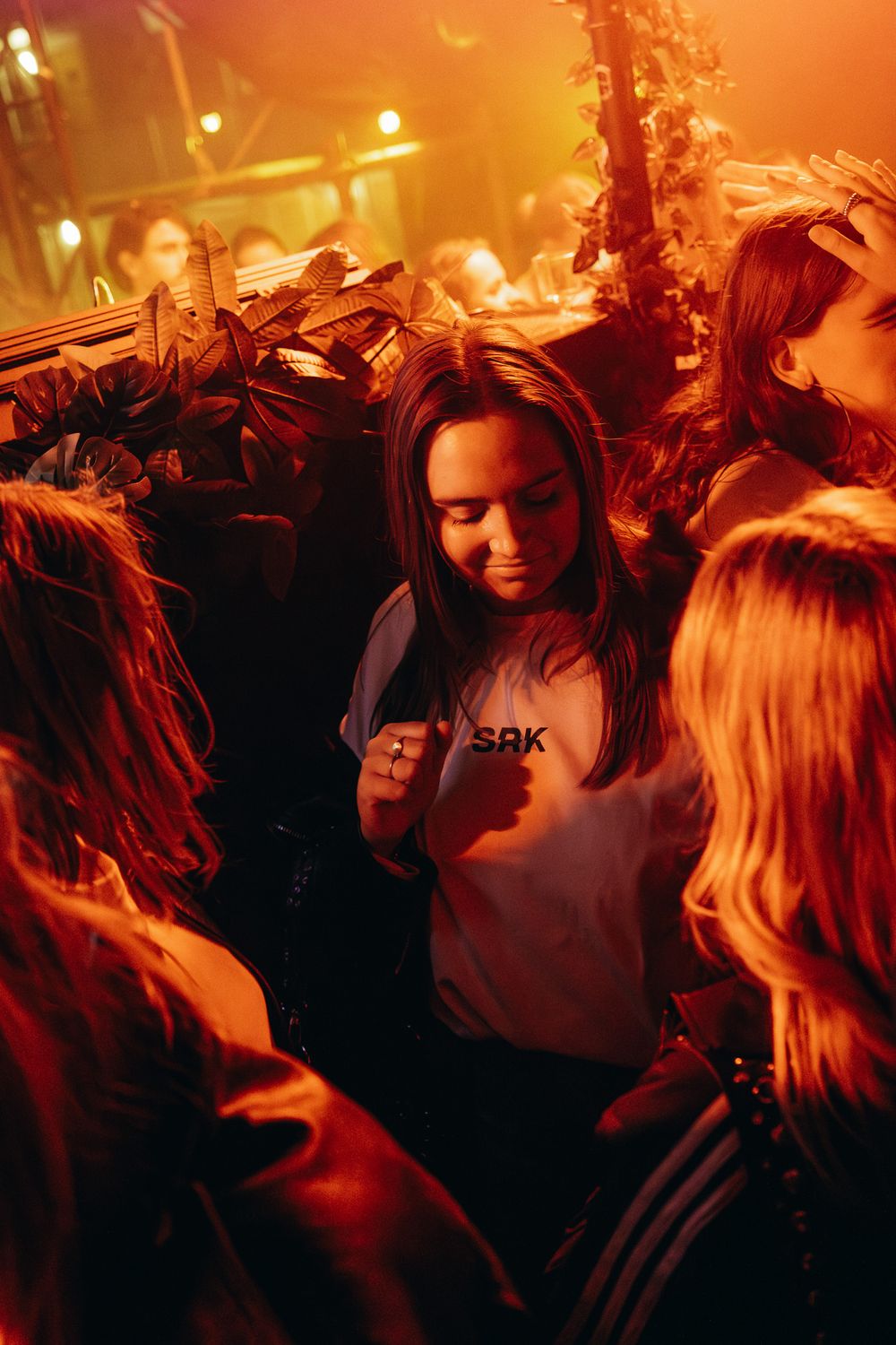 Young woman in white SBK shirt surrounded by friends at a vibrant nightclub with warm red lighting.