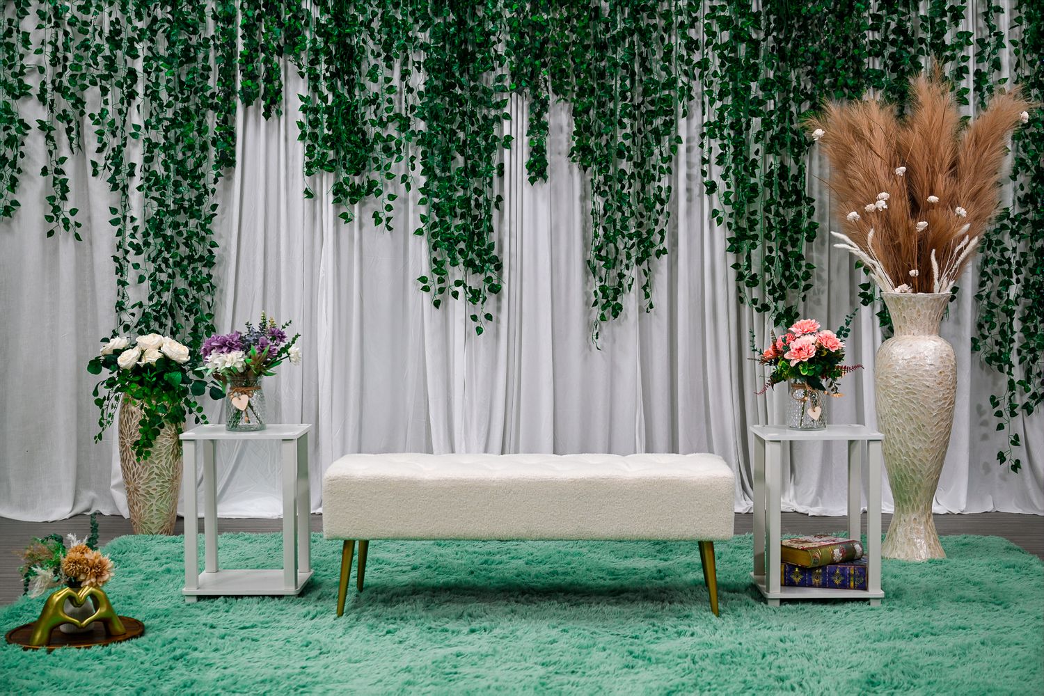 Elegant white bench with gold legs on green carpet, flanked by floral arrangements and hanging ivy backdrop.