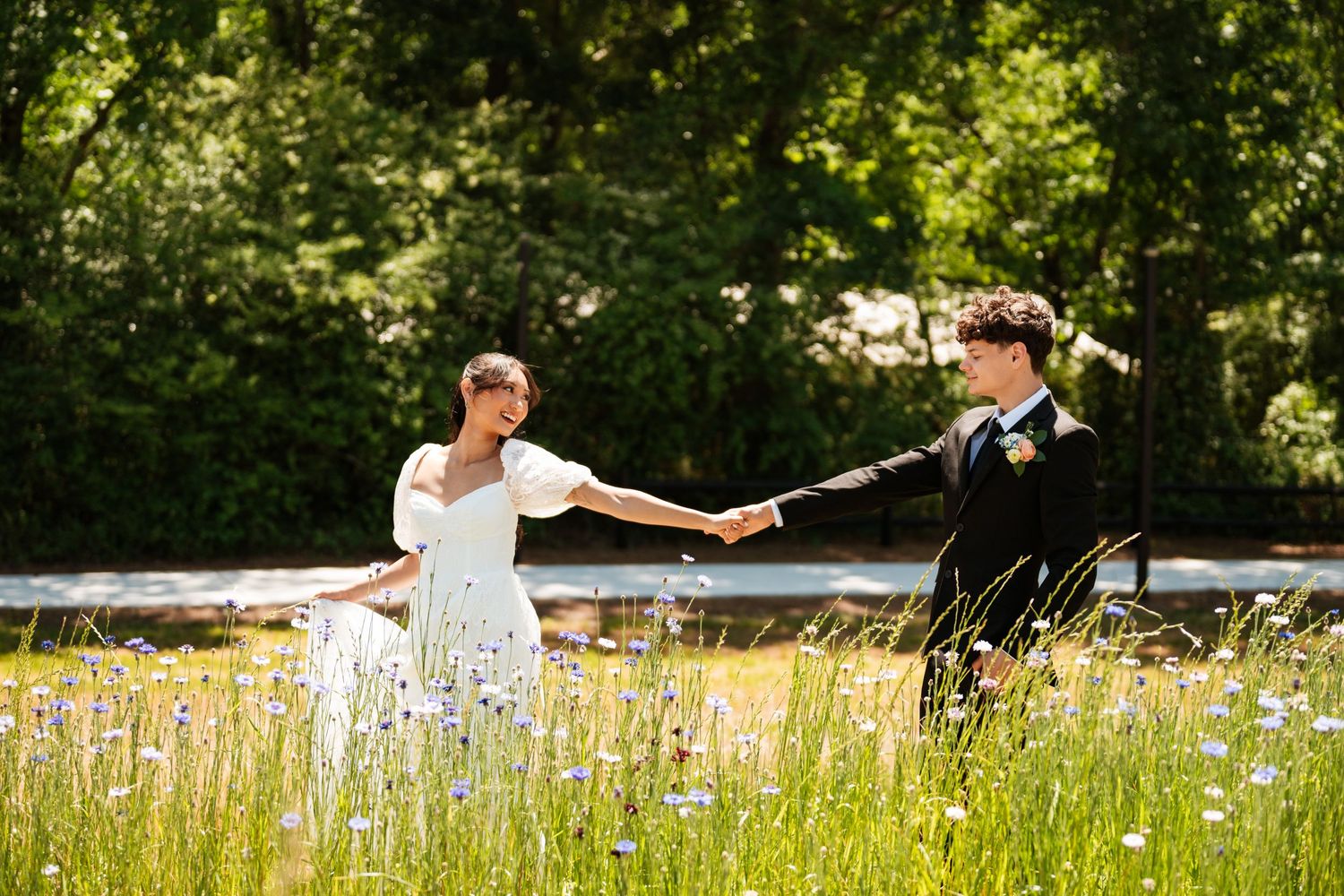 Bride and groom hold hands in a wildflower meadow with lush green trees in the background on their wedding day.