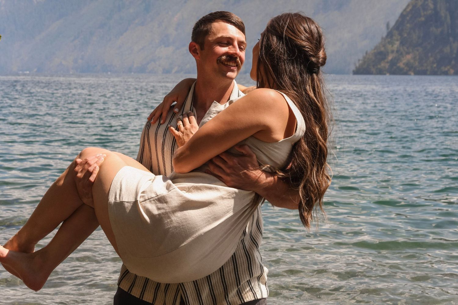 Man carrying woman in his arms, smiling by a scenic mountain lake on a sunny day.