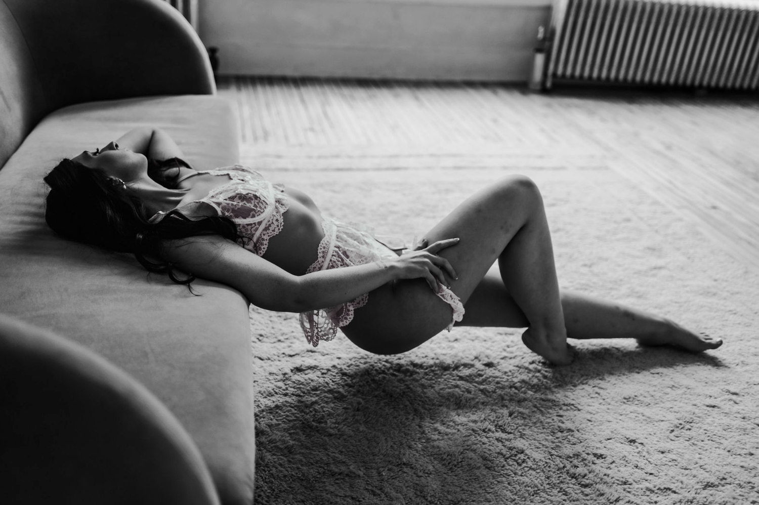 Black and white boudoir photo of a woman in lingerie reclining gracefully on a carpet beside a sofa.