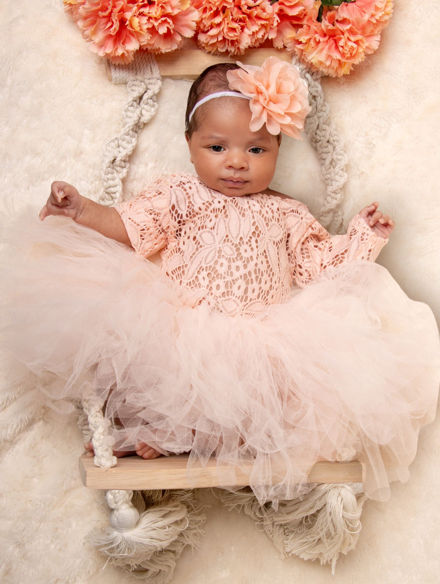 Newborn baby girl in peach lace dress and tutu with floral headband lying in decorative wooden swing.