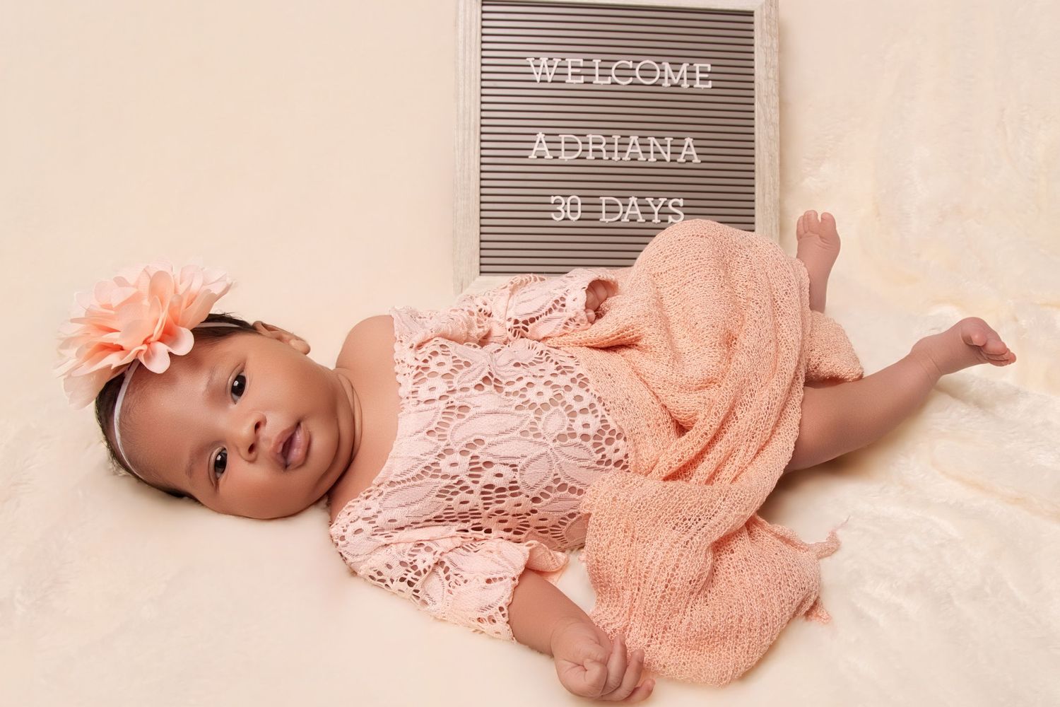 A baby girl named Adriana in a peach lace outfit and floral headband poses next to a welcome sign at 30 days old.