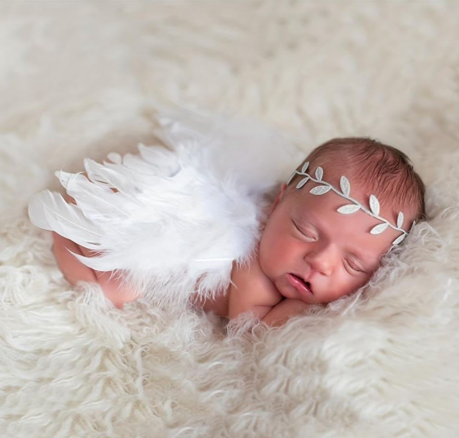 Newborn baby sleeping on fluffy white blanket wearing angel feather wings and silver leaf headband.