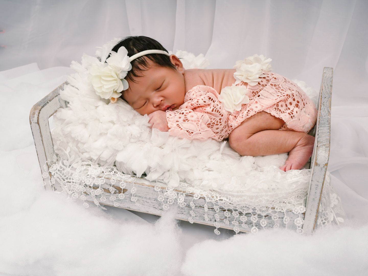 Newborn baby girl sleeping peacefully in a vintage white mini bed, wearing a pink lace outfit and white floral headband.
