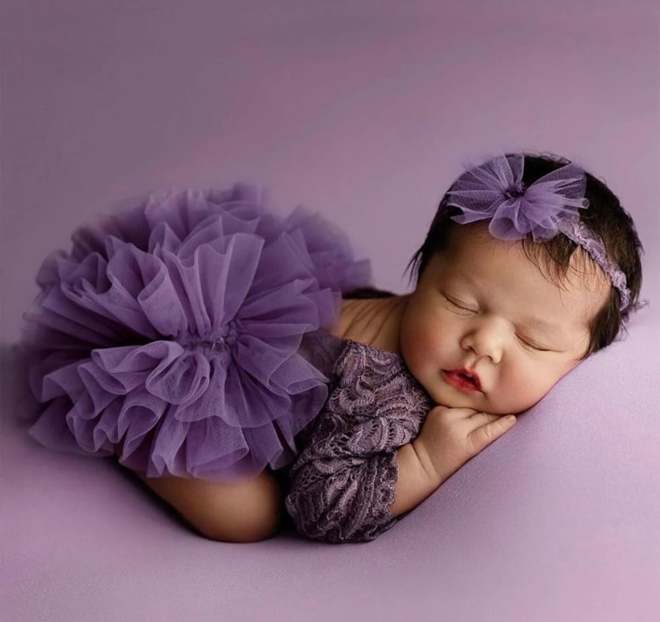Newborn baby girl sleeping peacefully in purple lace outfit and floral headband on lavender background.