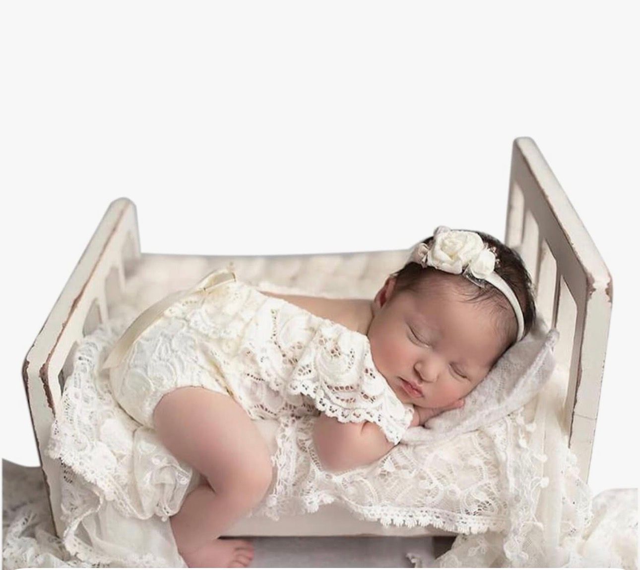 Newborn baby in lace romper and floral headband sleeping peacefully in a white vintage wooden crib.