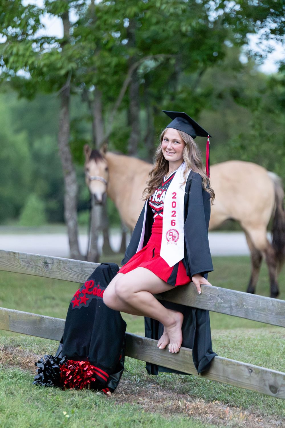 girl sitting on wood fence wearing cheer uniform, cap and gown. Letterman jacket next to her on the fence with her horse in the background