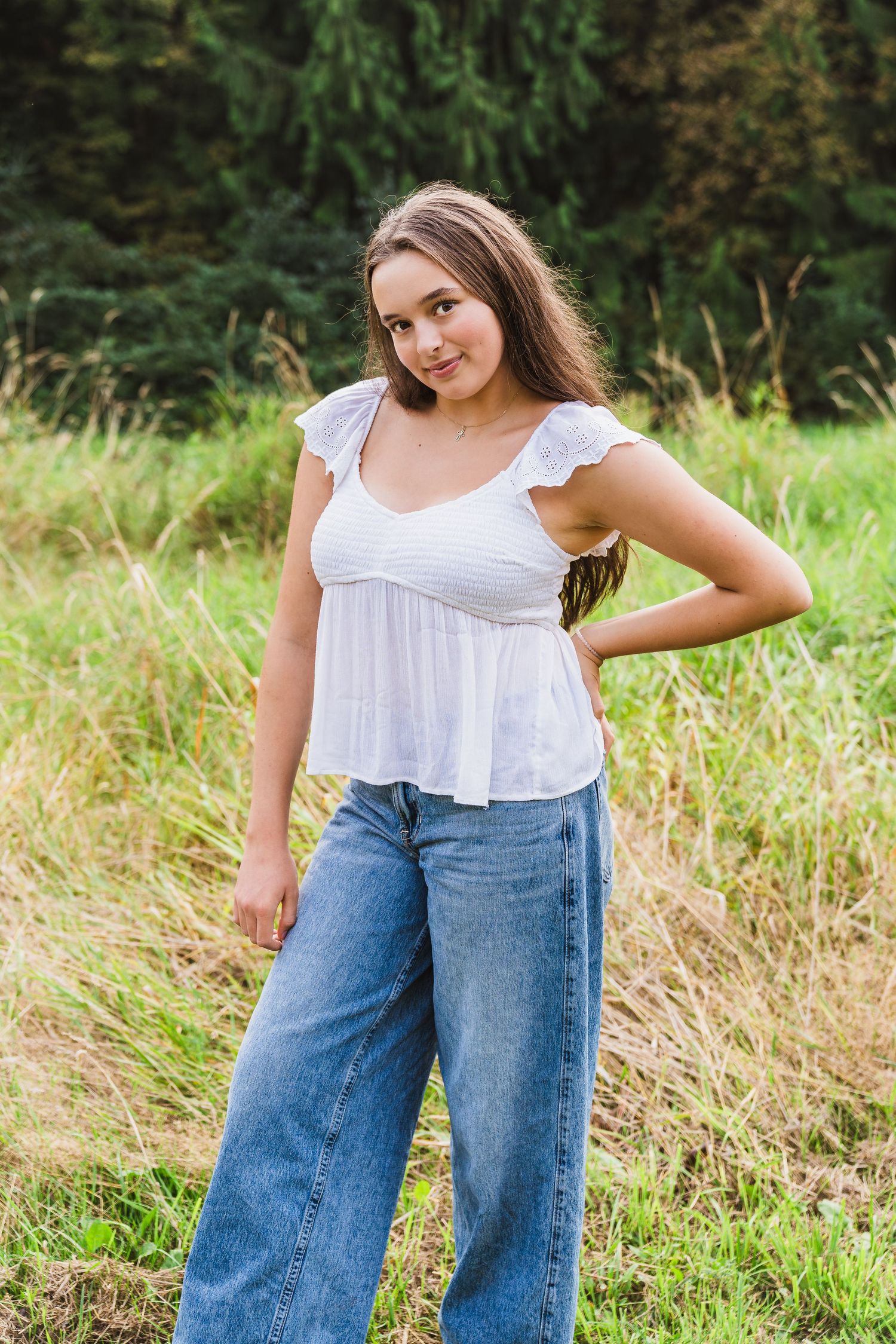 Young woman in white ruffled top and wide-leg jeans posing outdoors in a grassy field with trees behind her.