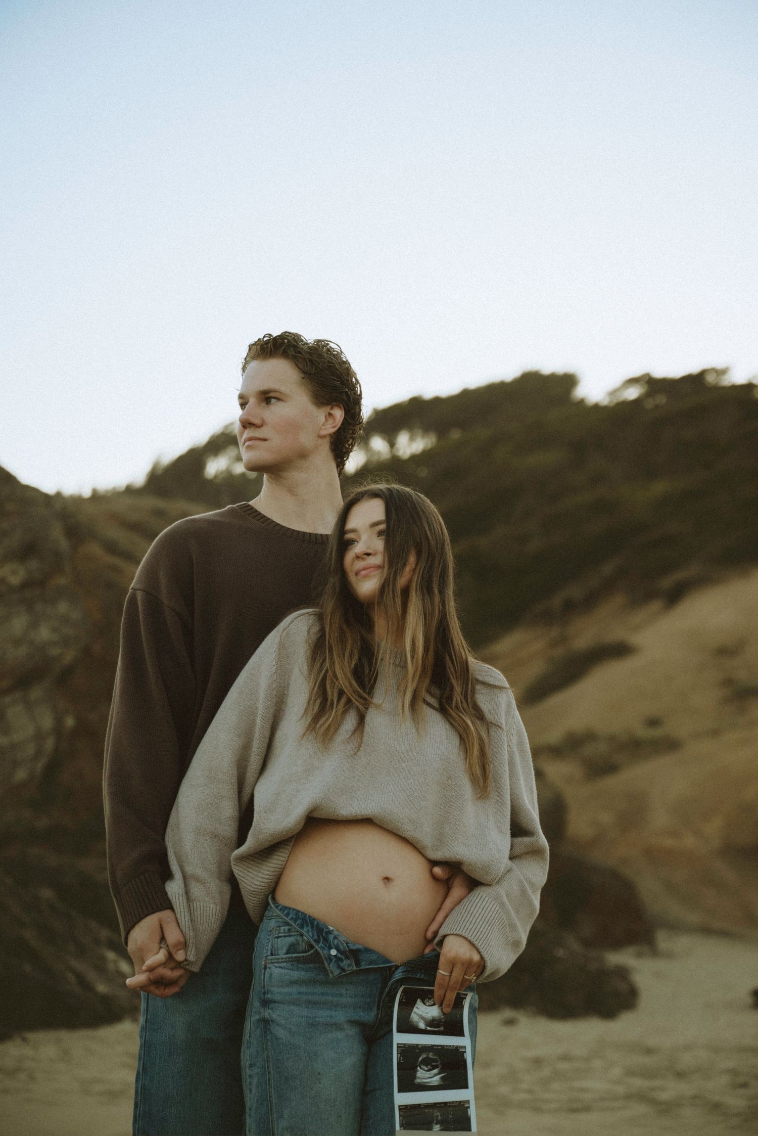 Pregnant couple holding ultrasound photos on a beach, woman showing baby bump in casual sweater and jeans outdoors.