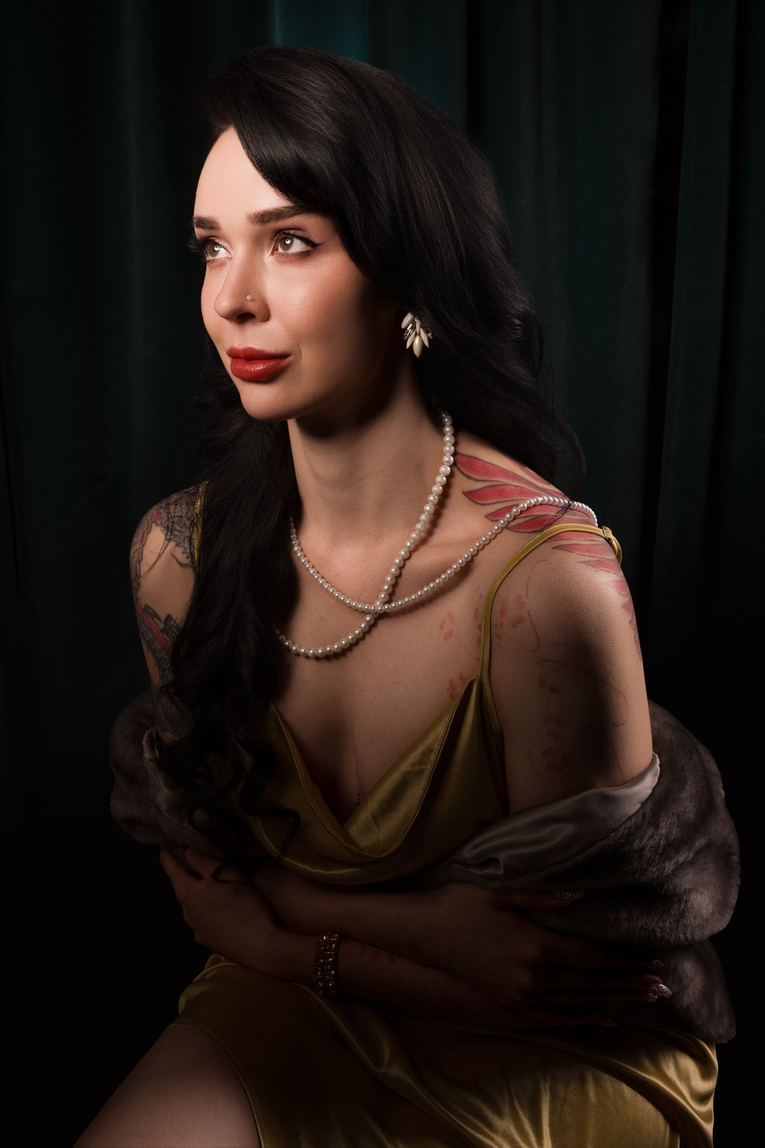 Dark-haired woman in gold satin dress with pearl necklace and floral shoulder tattoo poses against green curtain backdrop.