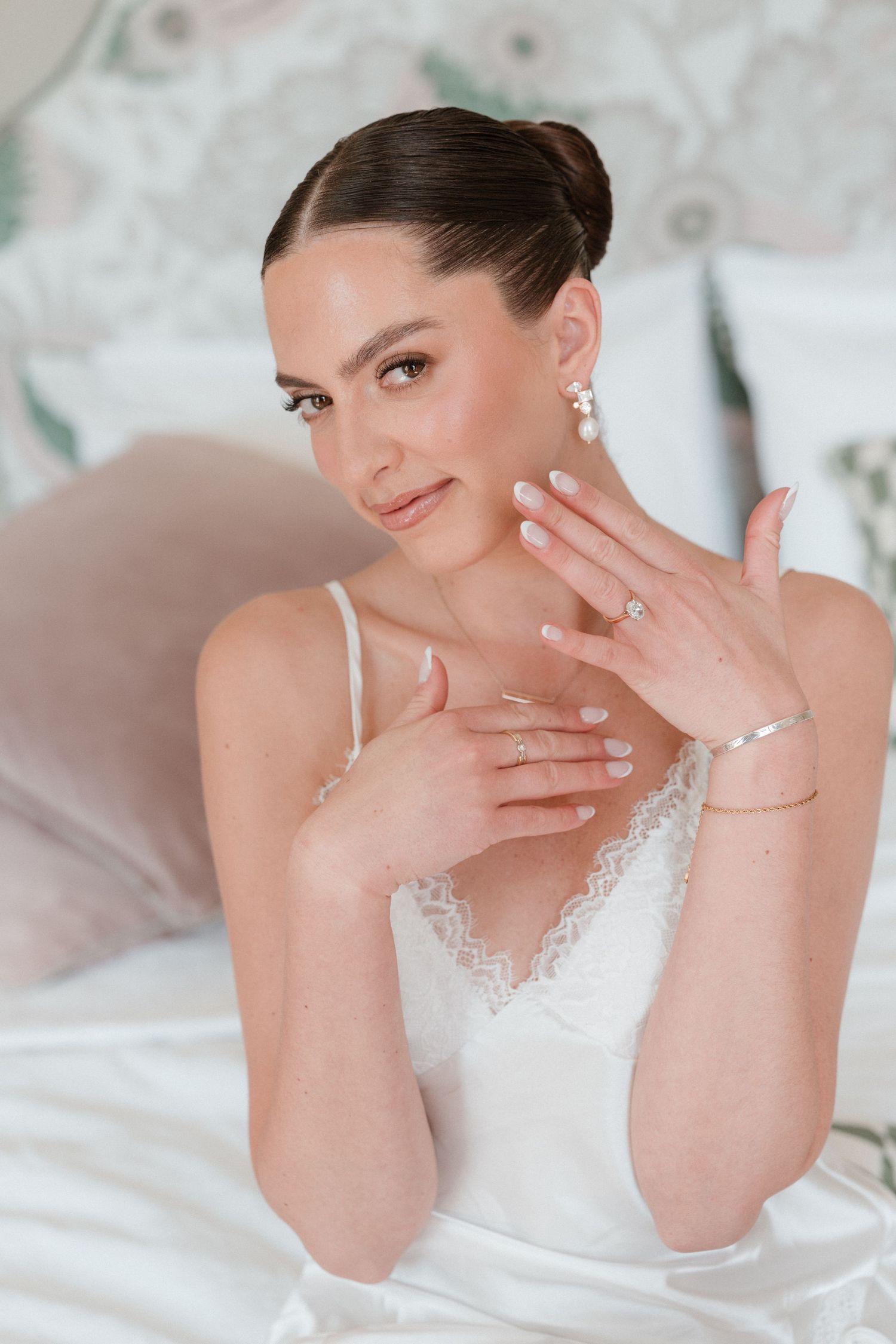 Elegant bride in white lace dress with pearl earrings, bracelet, and ring, posing gracefully on a bed.