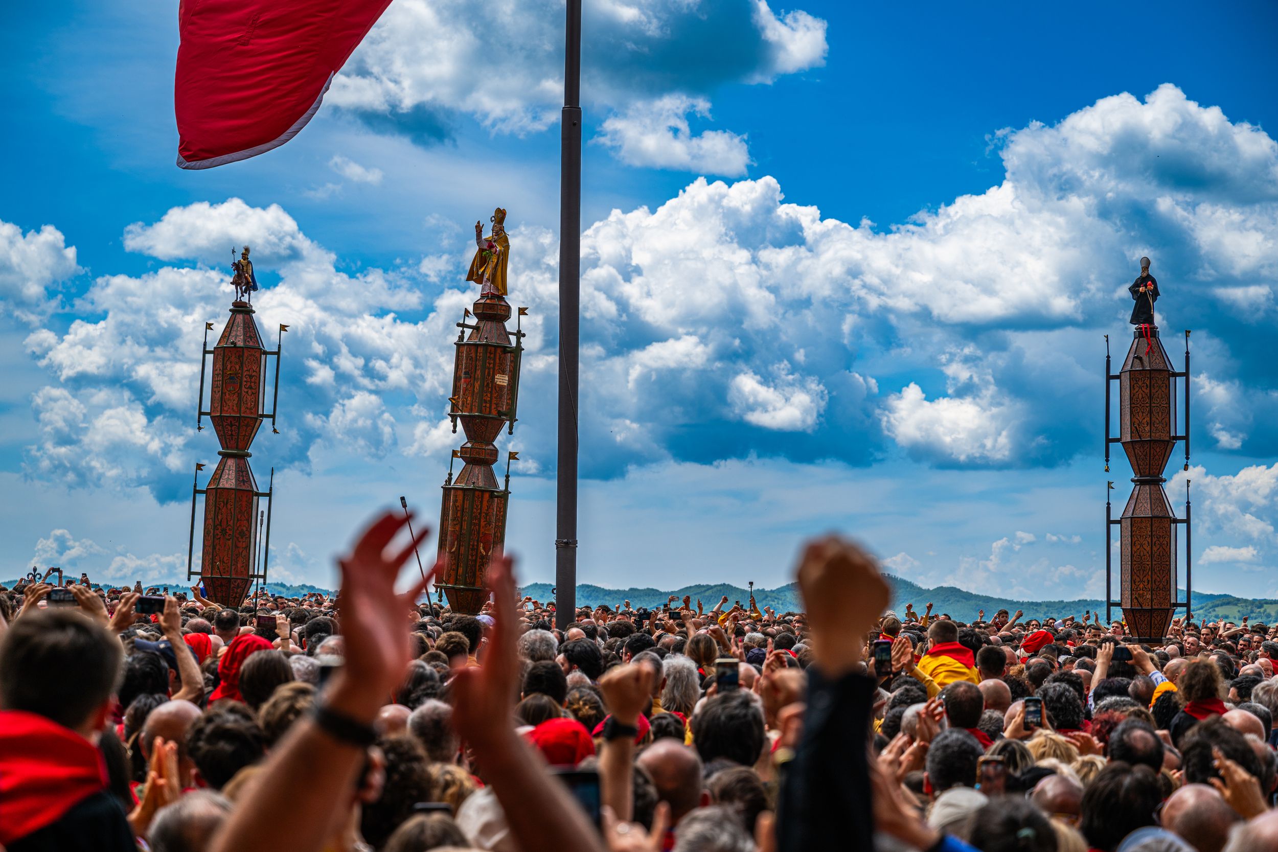 Large crowd at outdoor rally with raised fists, golden statues on poles, red flag, and dramatic blue sky with clouds.