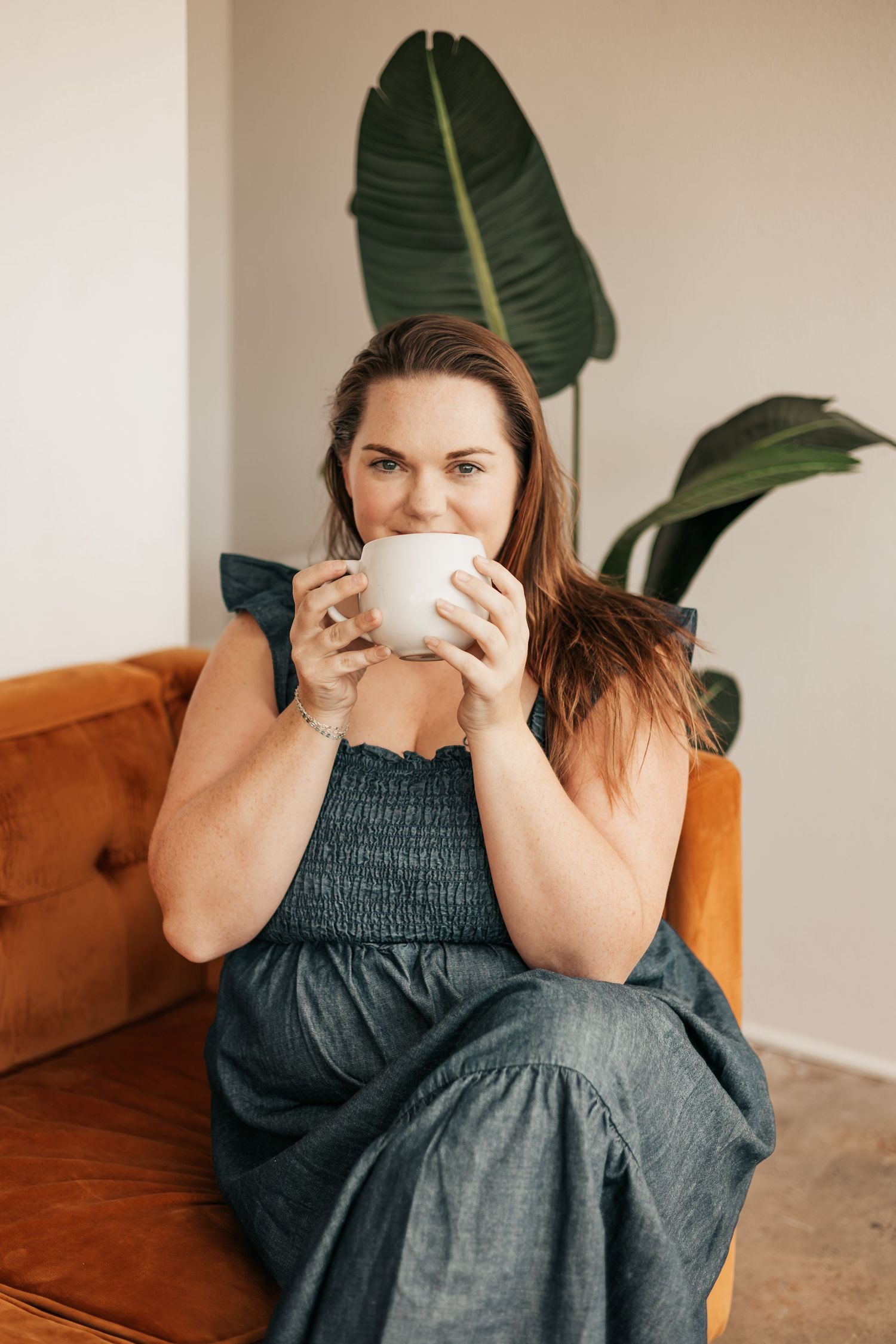 Woman in blue dress sipping from white mug on orange sofa with tropical plant in bright modern interior | Photography Business Coach