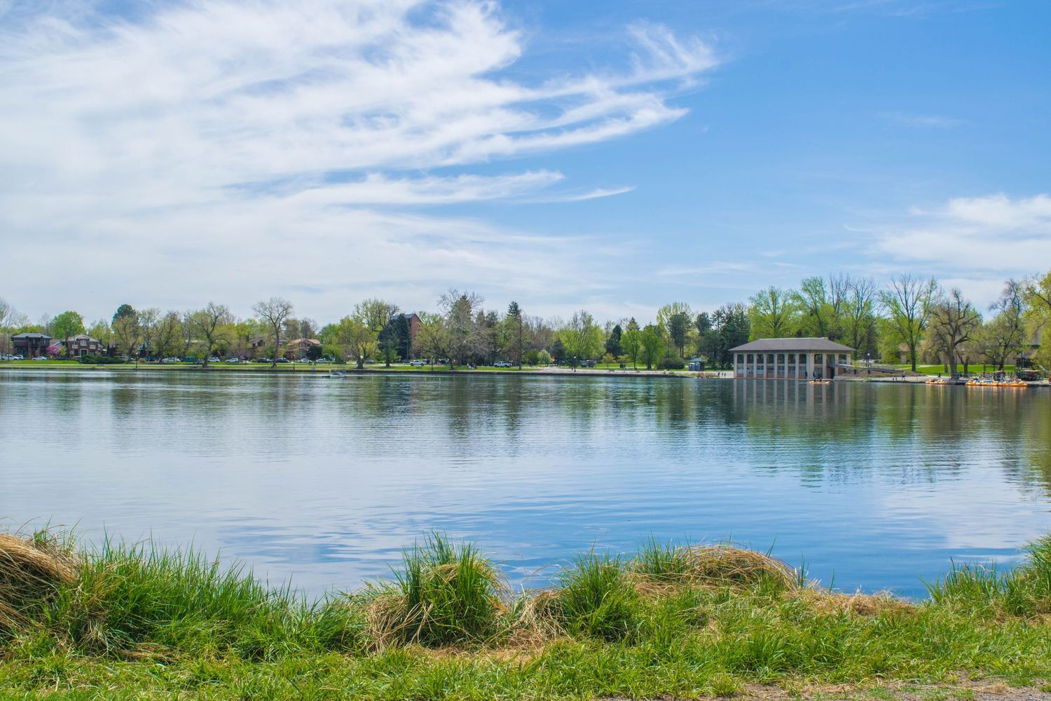 Serene lake in a lush green park with a gazebo, blue sky, and wispy clouds reflected on calm water.