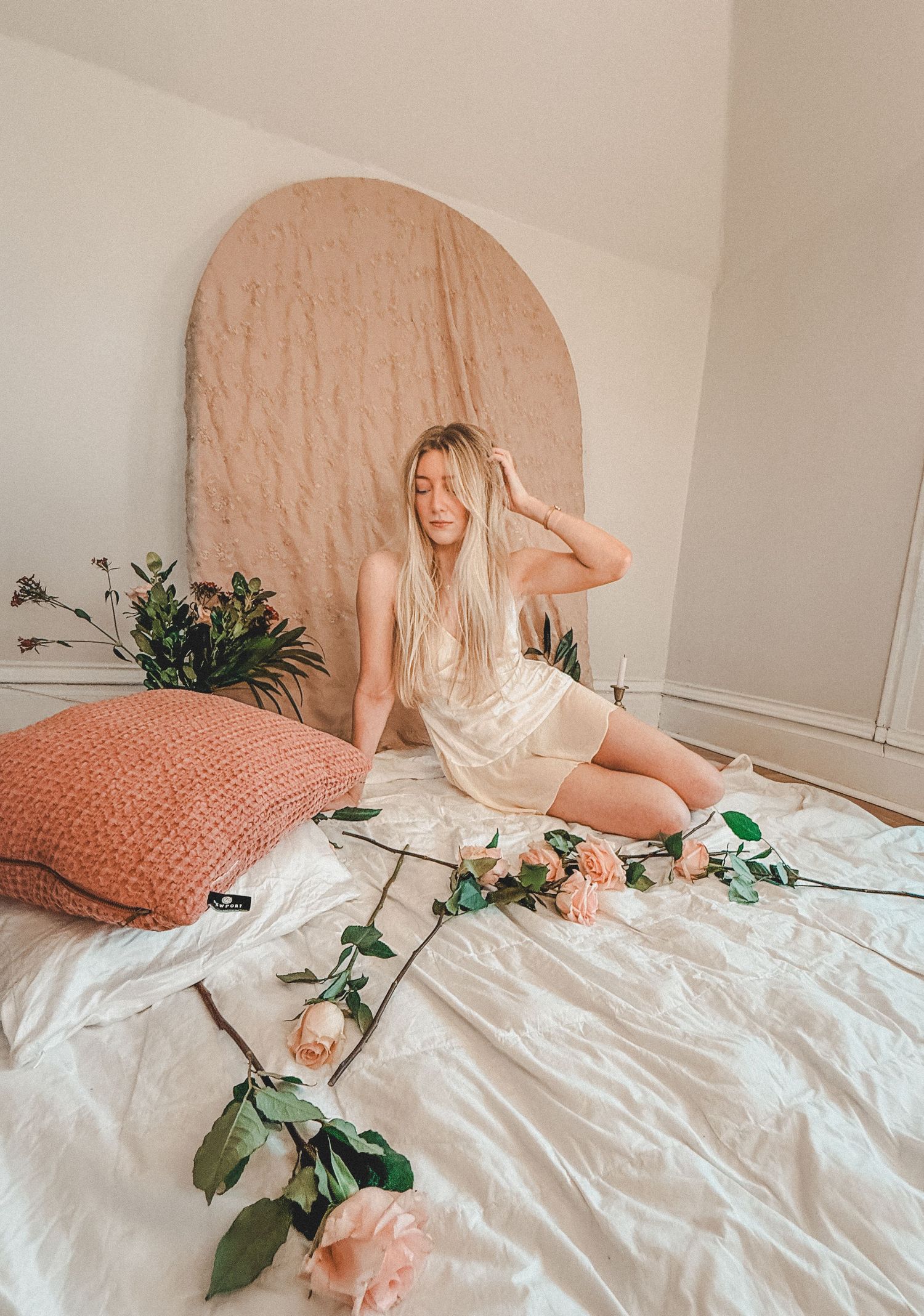 Blonde woman in cream satin loungewear sitting on white bedding surrounded by pink roses and a blush arch backdrop.