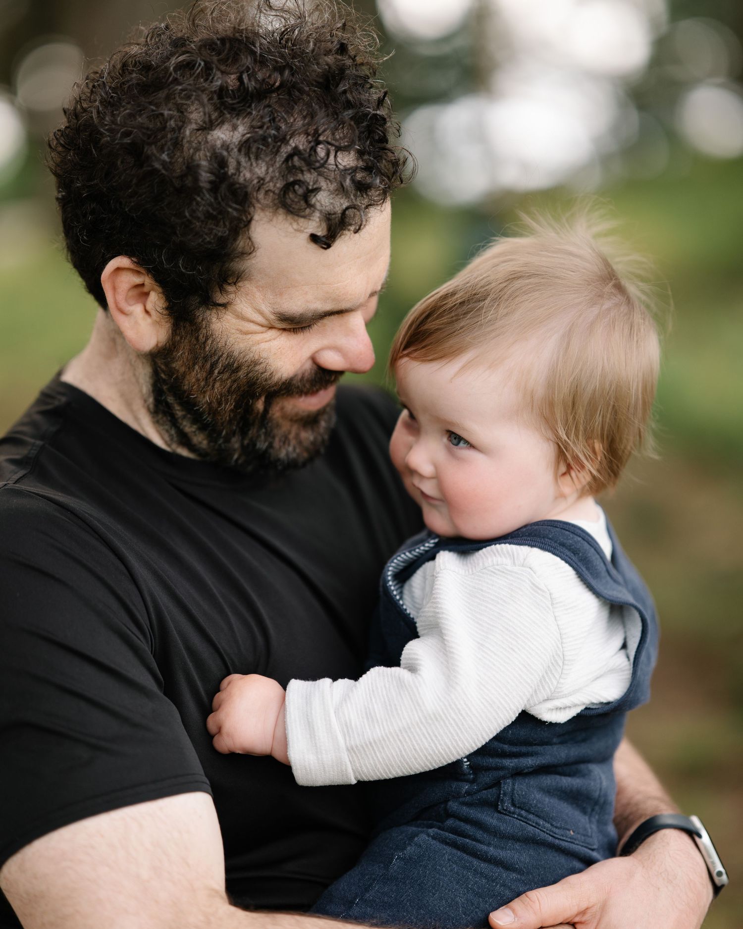 A bearded man with curly hair holds a smiling baby outdoors, sharing a tender father-child moment.