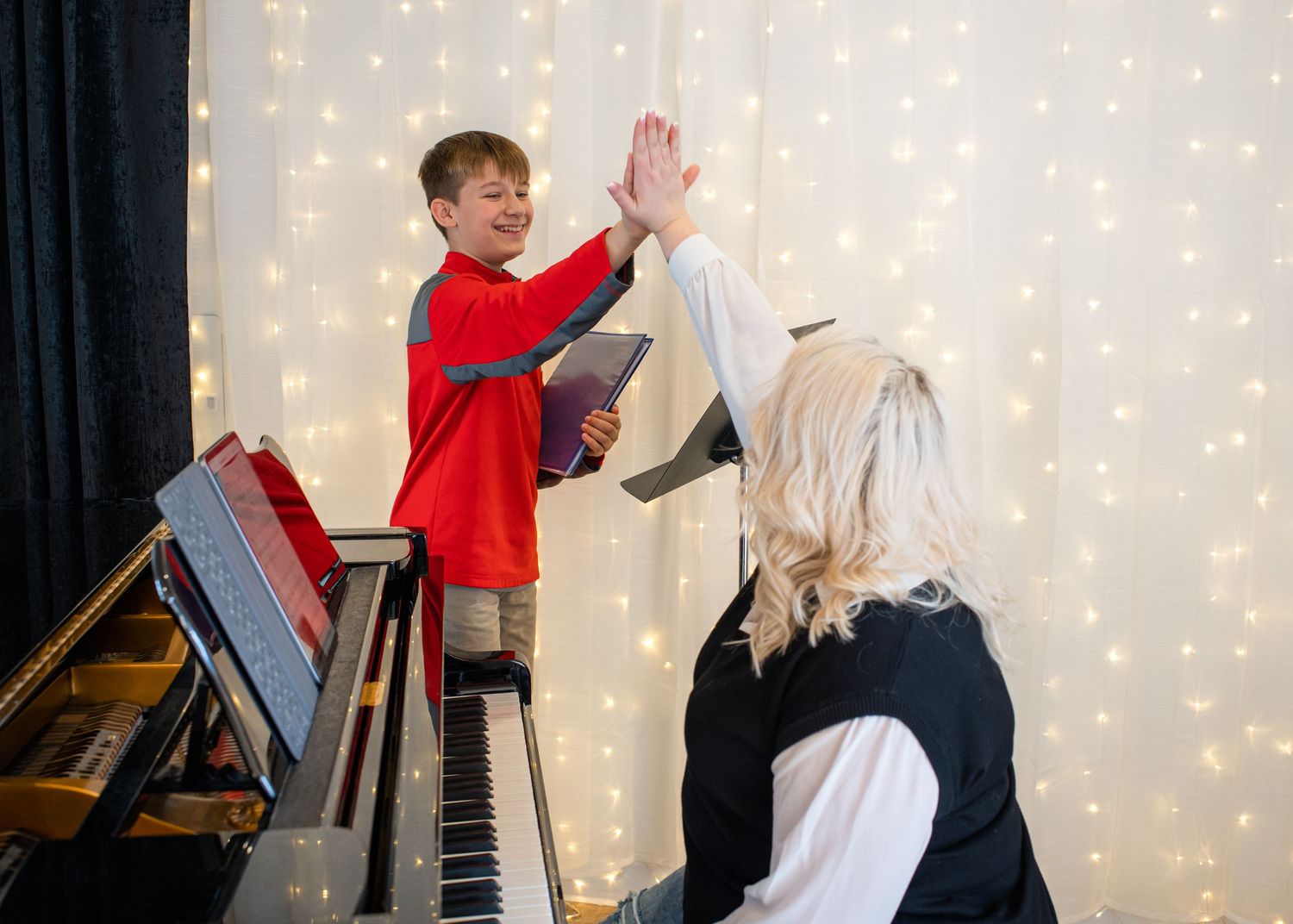 A young boy in a red jacket high-fives his blonde piano teacher near a grand piano with fairy lights backdrop.