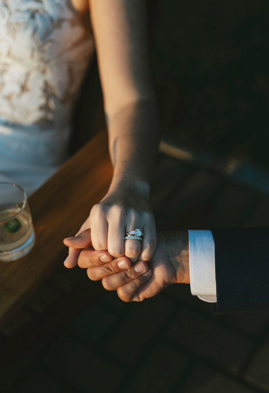 Bride and groom holding hands at wedding, showcasing elegant diamond engagement ring, warm golden light.