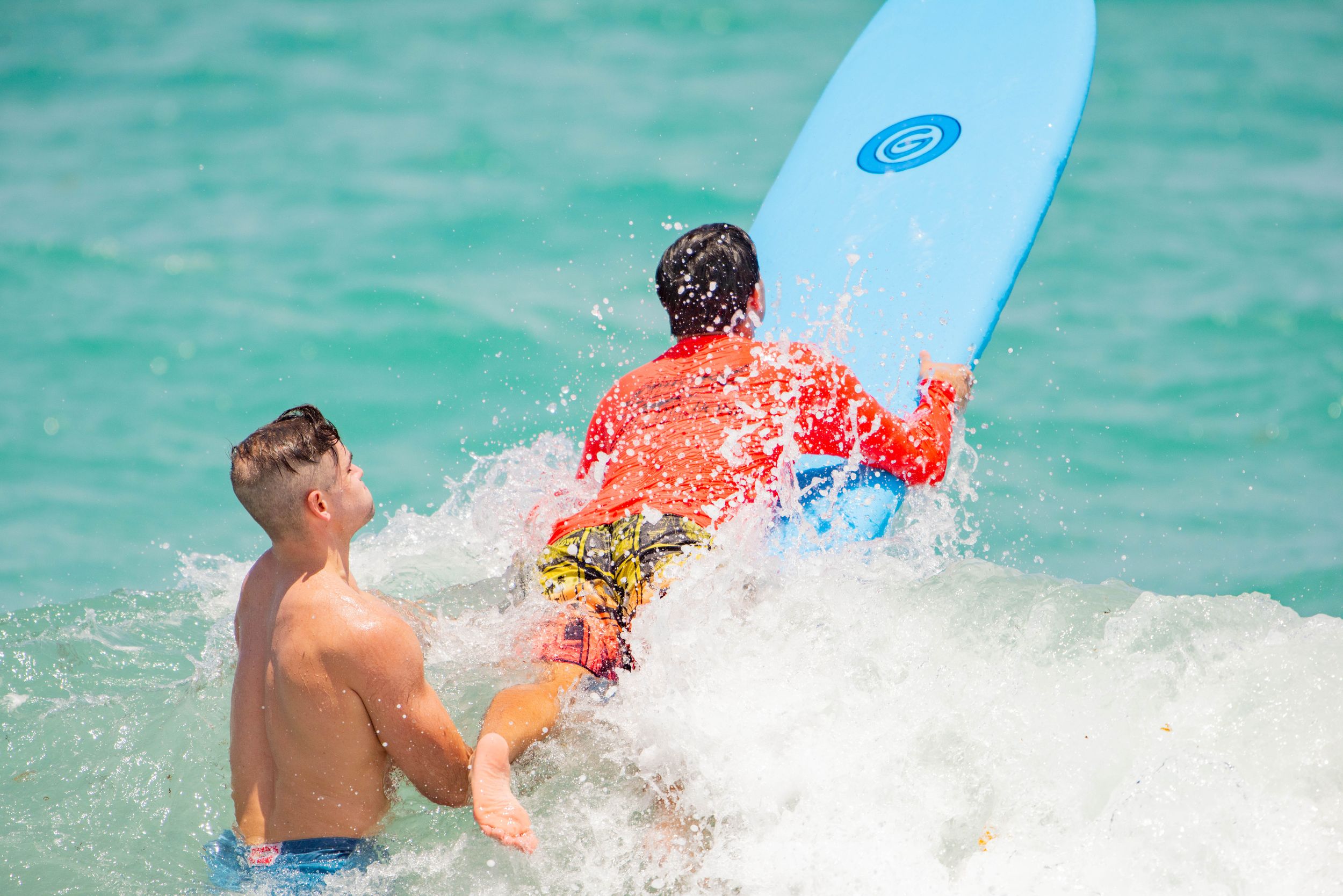 Two boys playing in ocean waves with a blue surfboard on a sunny tropical beach day.