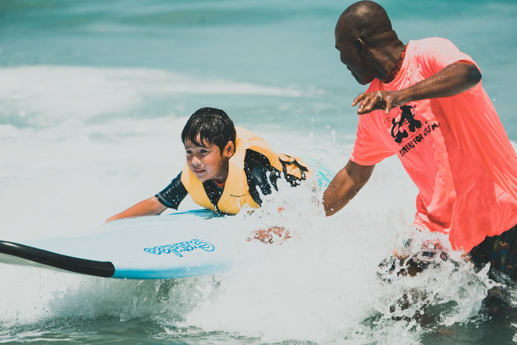 Surf instructor in pink shirt teaching young child to surf on a longboard in the ocean.