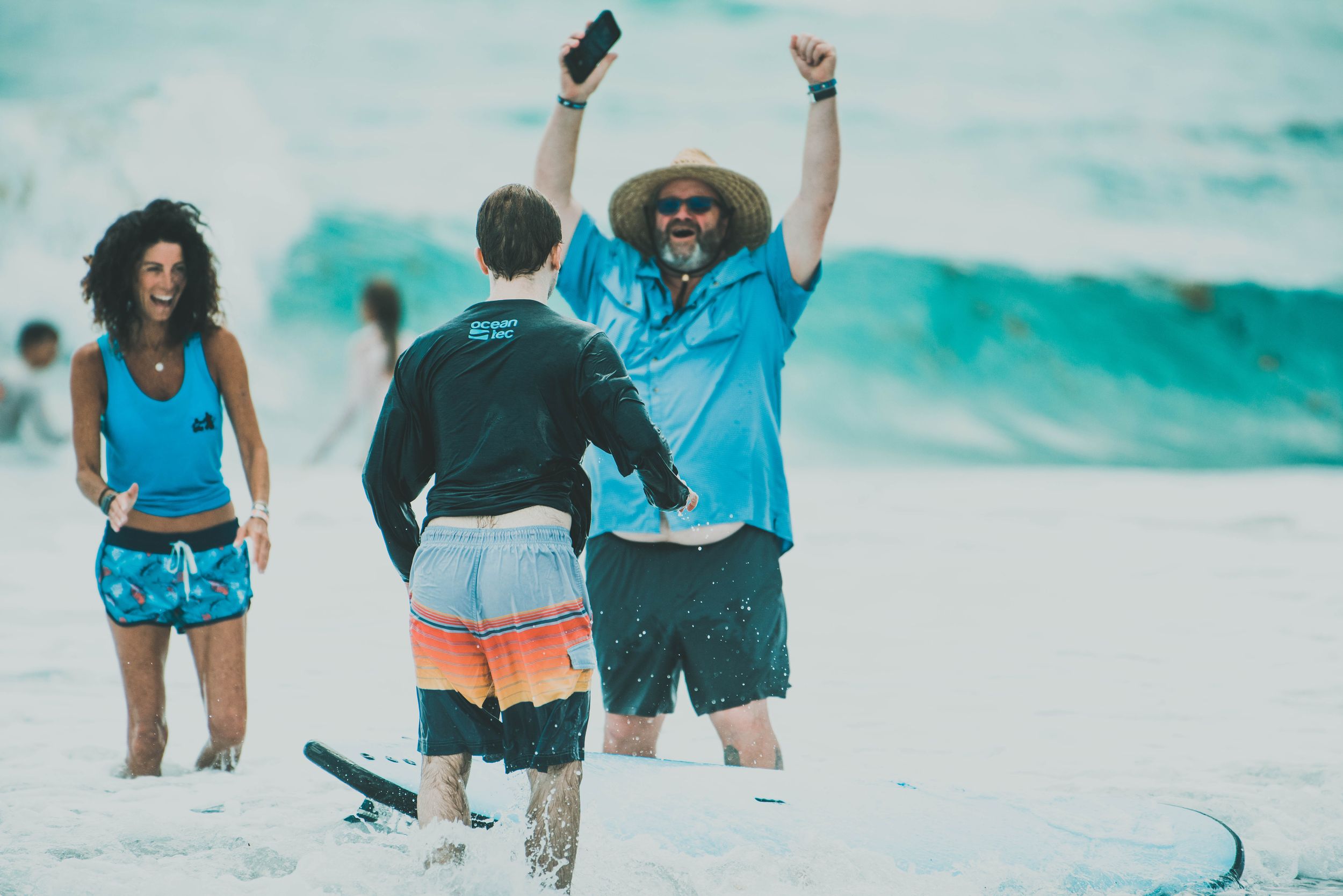 A surf instructor celebrates with raised fists with a student on a sunny beach with waves in the background.