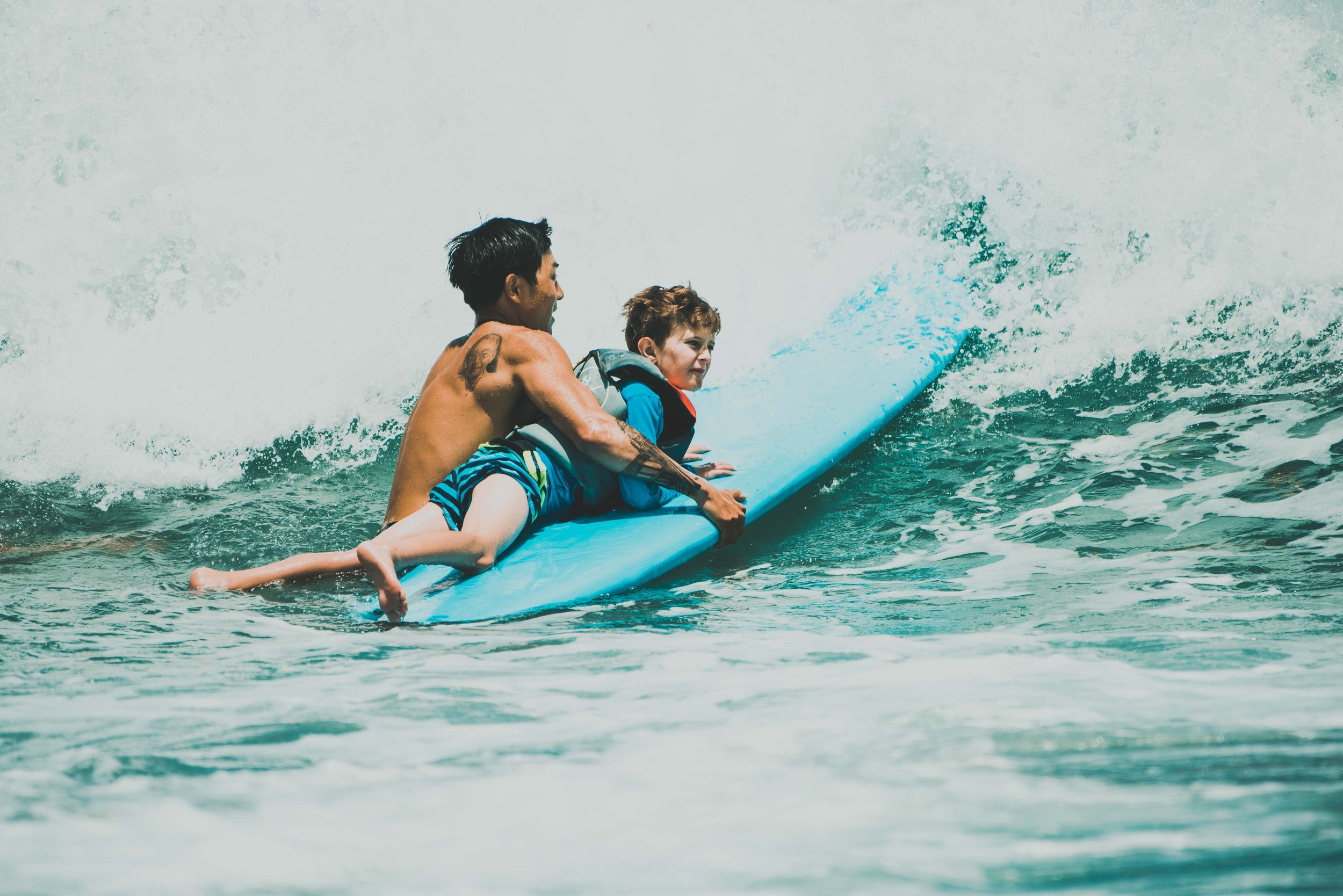 Two people riding a blue surfboard together on ocean waves, with an adult helping a child learn to surf.