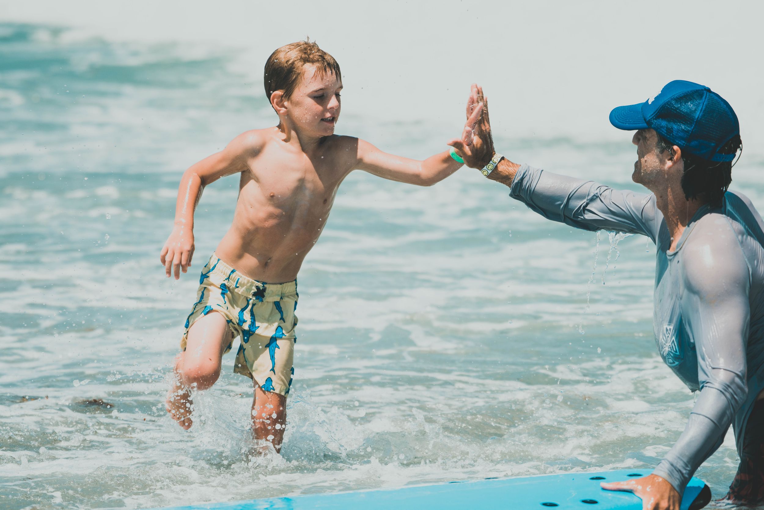 A young boy high-fives an adult instructor while learning to surf in the ocean waves on a sunny day.