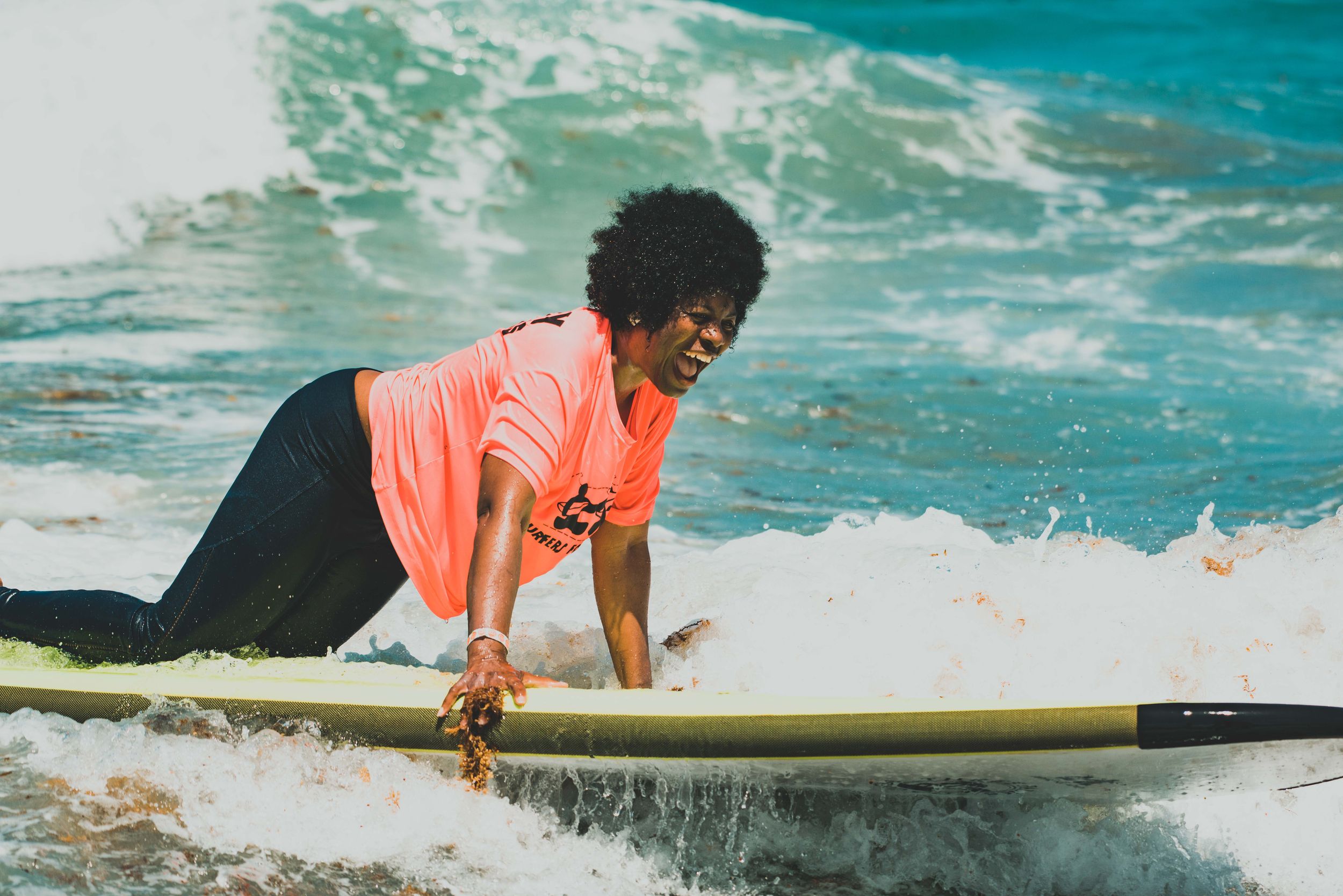 A smiling Black woman with an afro rides a surfboard through ocean waves wearing a coral pink shirt.