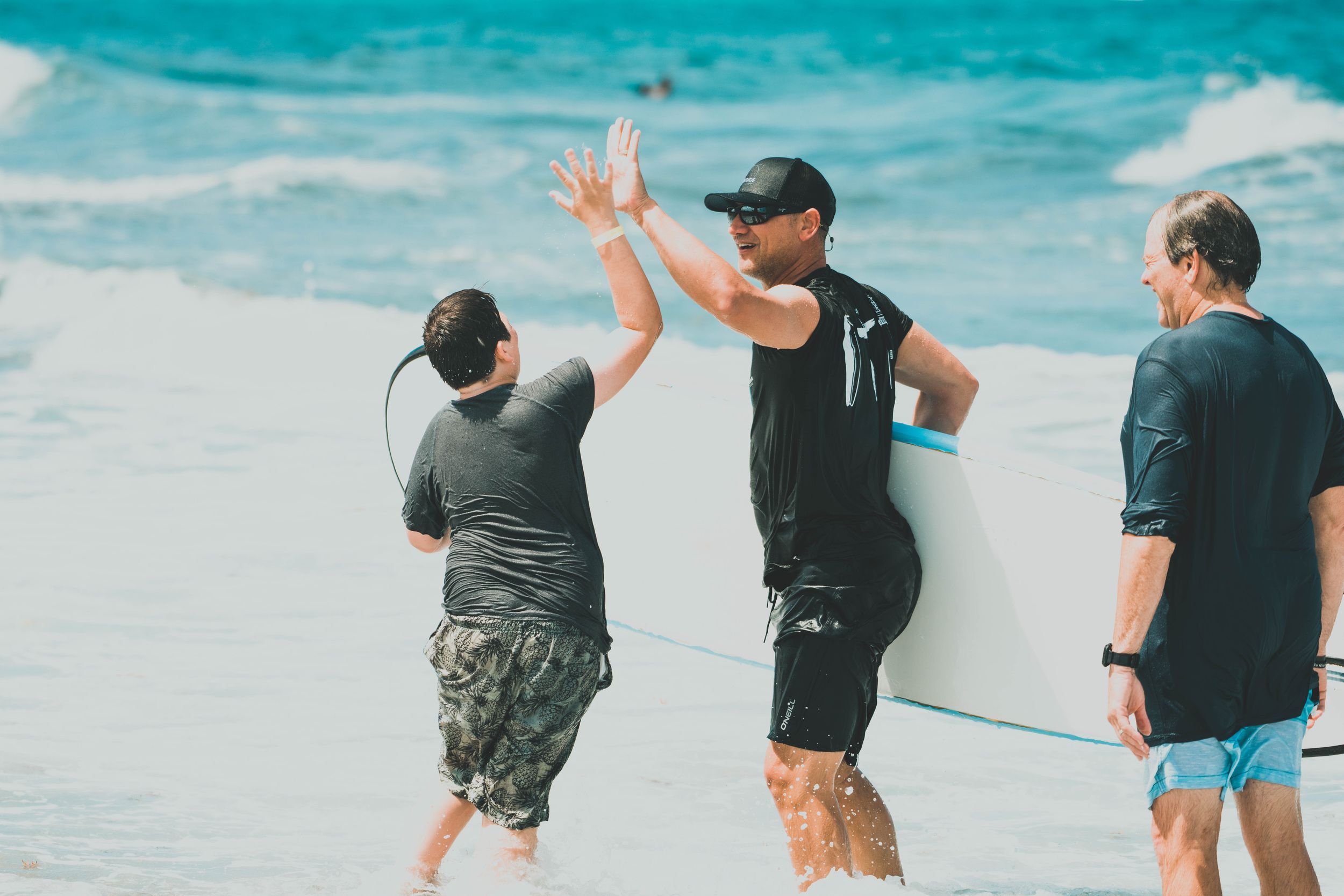 Two men high-fiving on a sunny beach while a third person holds a white surfboard nearby.