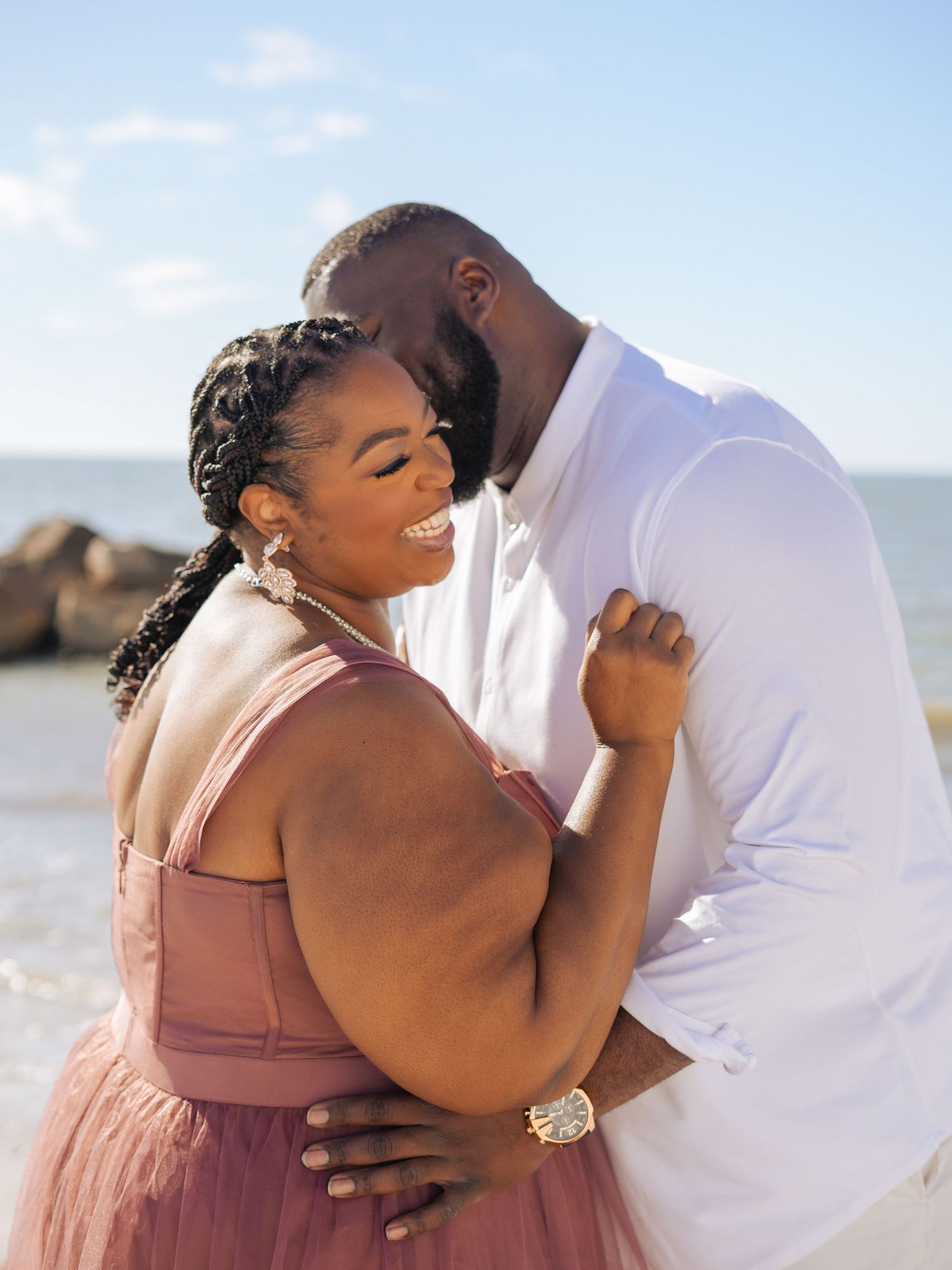 Couple posing together on a sunny beach, woman in pink tulle dress and man in white outfit, with rocky shoreline behind them.