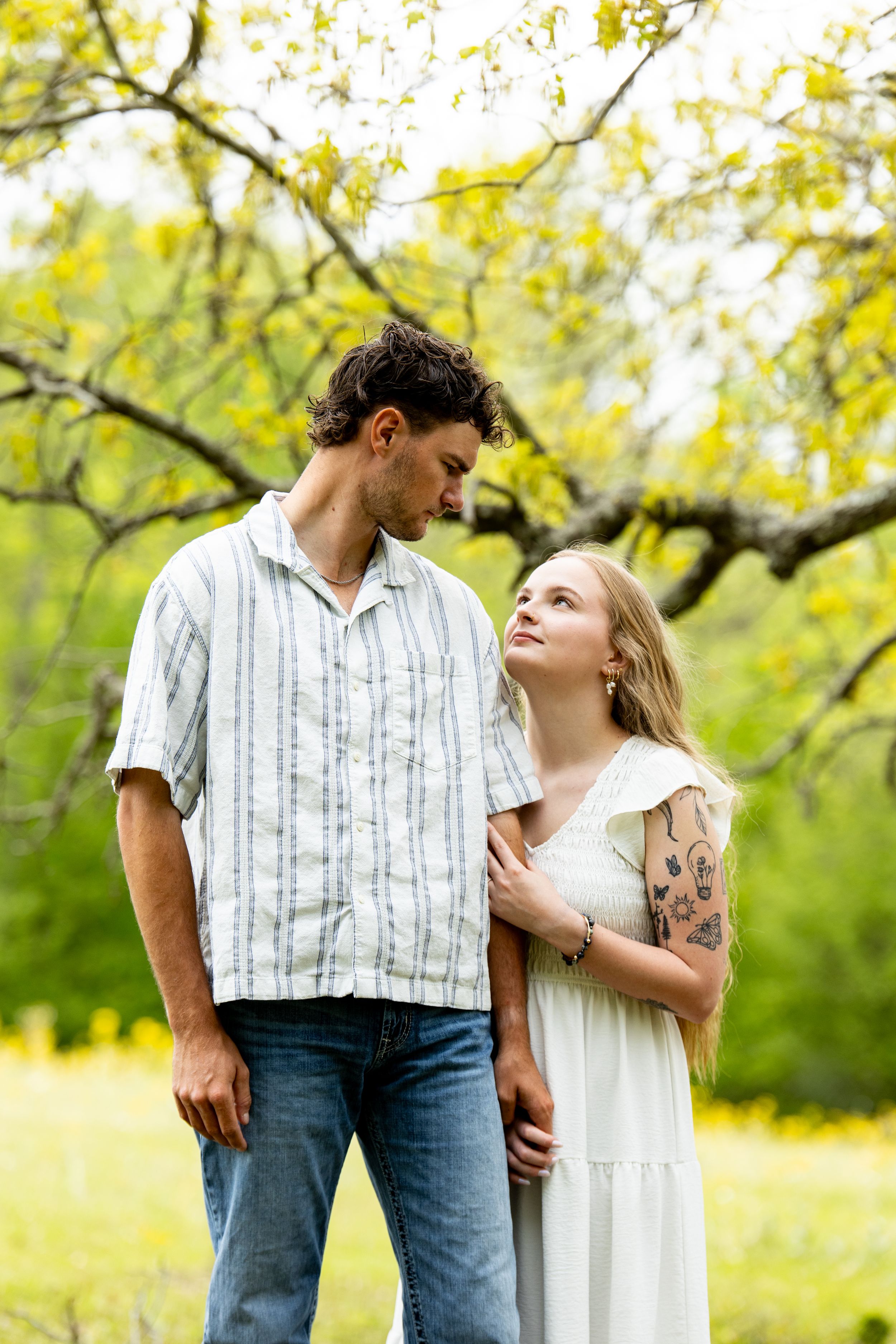 Couple standing together in a blooming spring garden, woman with tattoos in white dress gazing up at man in striped shirt.