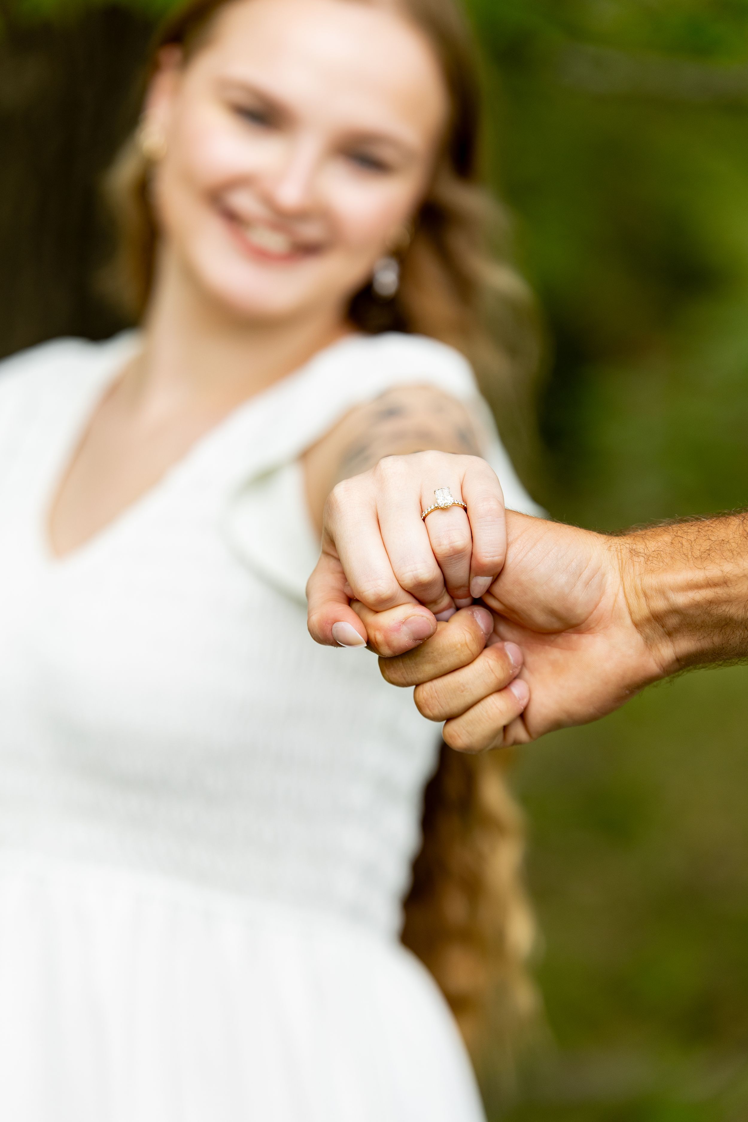 Smiling bride in white dress showing off her engagement ring while holding hands with groom outdoors.