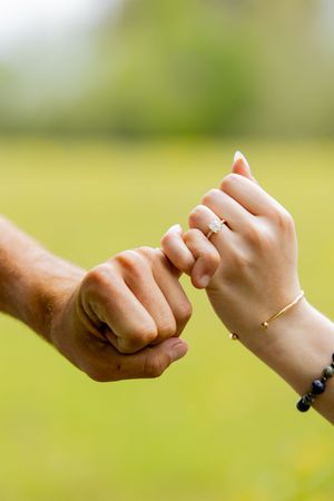 Couple linking pinky fingers outdoors, woman wearing diamond engagement ring and gold bracelet on green background.