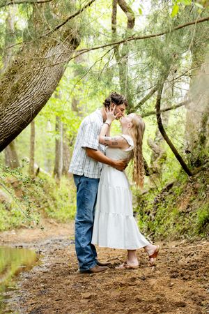 Couple sharing a romantic kiss on a forest path surrounded by lush green trees and a small stream.