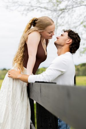 A romantic couple shares an intimate moment leaning over a dark wooden fence in a lush green outdoor setting.