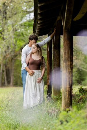 Romantic couple embracing near rustic wooden structure surrounded by lush greenery in a serene outdoor setting.