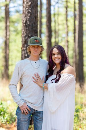 Couple posing in a pine forest, man in green cap and jeans, woman in white off-shoulder dress with purple hair.