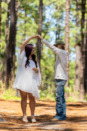 Couple dancing together on a forest path, woman in white dress being twirled by man in jeans and green cap.