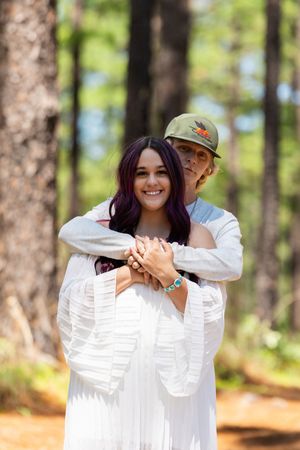 Couple embracing in a forest, woman in white dress with purple hair, man in green cap, smiling outdoors.