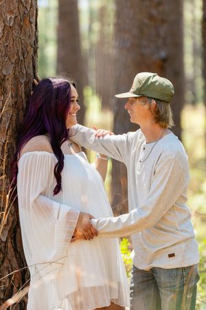 A couple shares a tender moment in a sunlit forest, the woman in white with purple hair leaning against a pine tree.