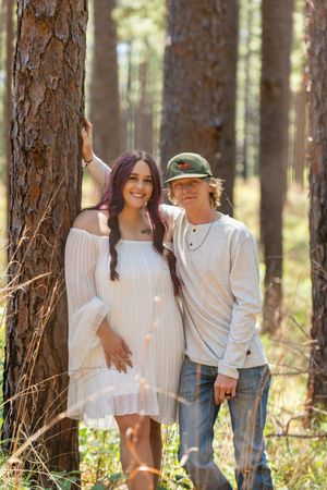 Couple posing in a sunlit pine forest, woman in white dress with purple hair, man in green hat and jeans.