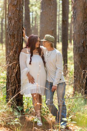 A couple shares a tender moment leaning against a pine tree in a sunlit forest, woman in white dress, man in cap.