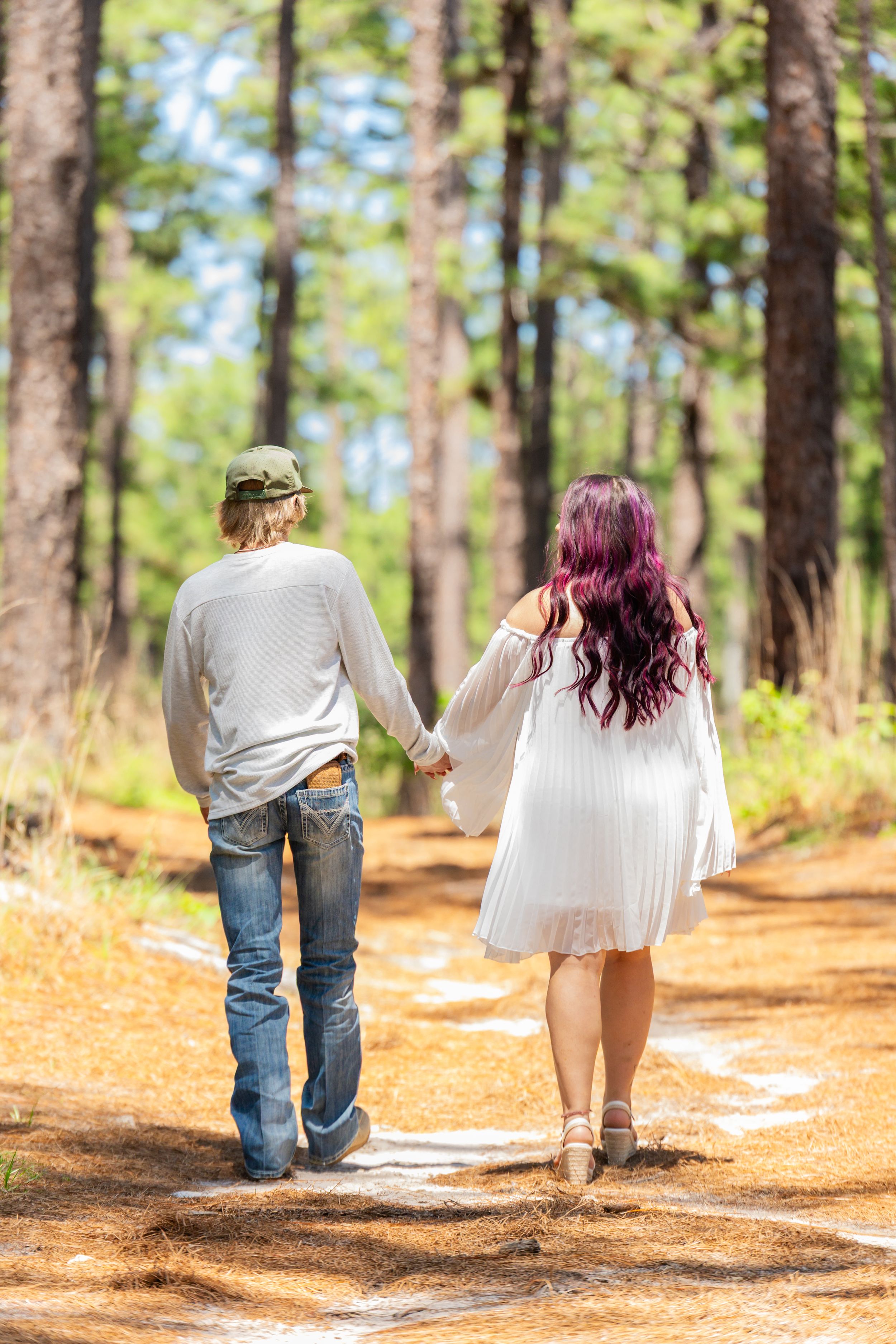 Couple holding hands walking on a forest path, woman in white dress with purple hair, man in jeans and cap.