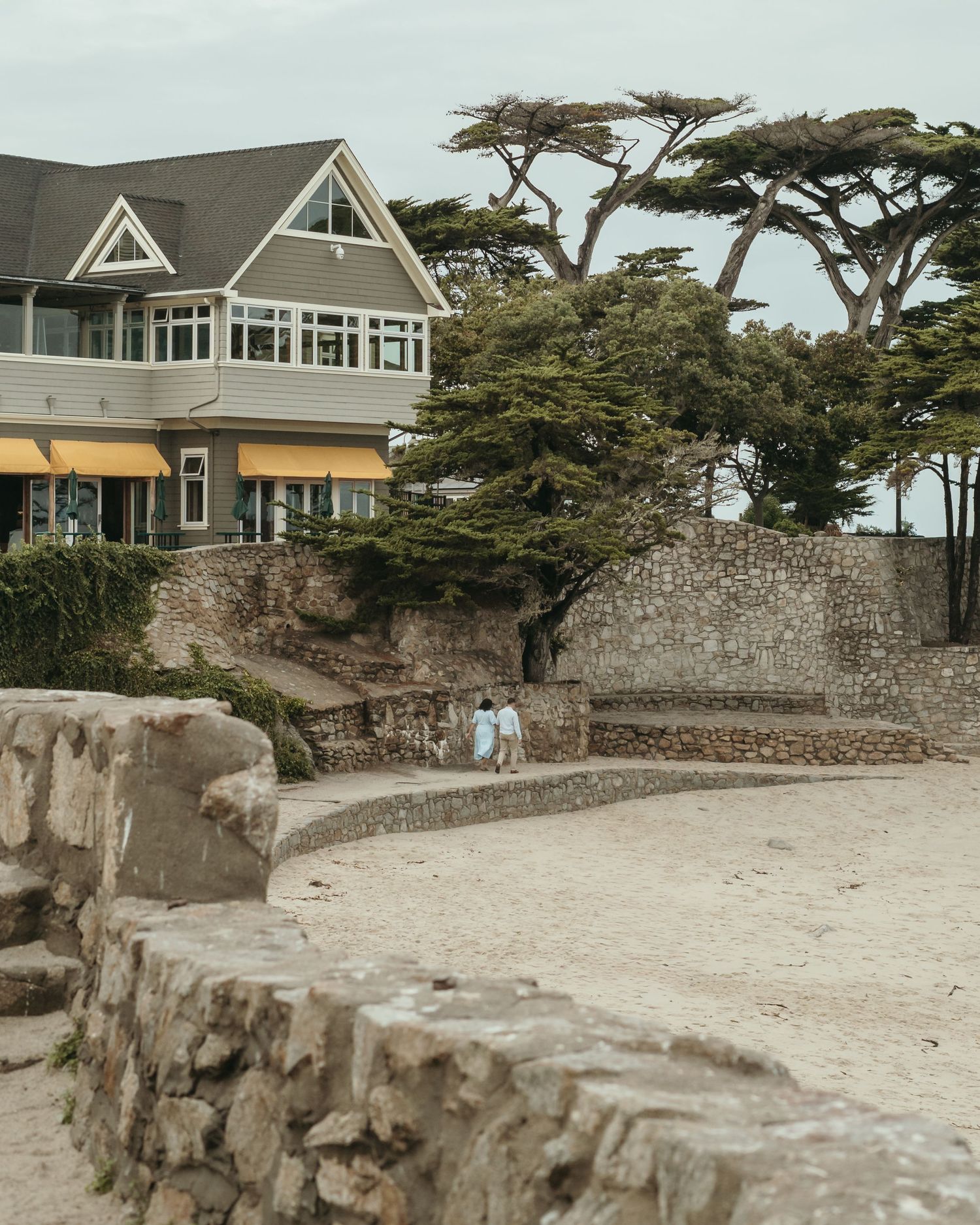 Beachside house with yellow awnings and stone walls near sandy shore with cypress trees in background.
