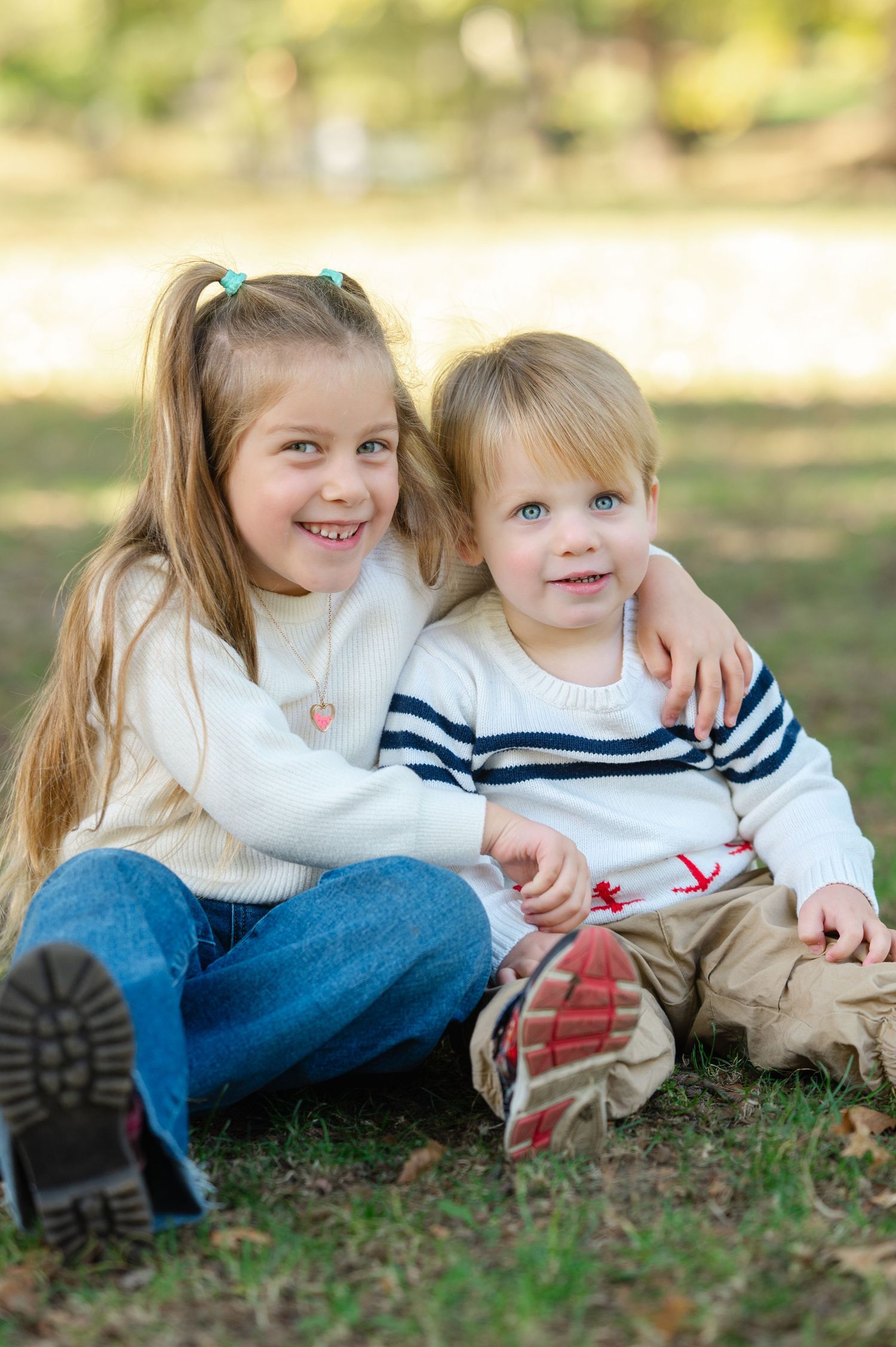 Two young siblings sitting together on grass in a park, smiling, wearing cozy sweaters in an outdoor portrait.