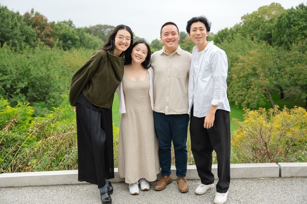 Four young Asian friends smiling together for a group photo in front of a lush green landscape.