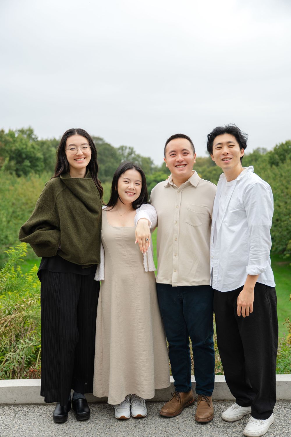 Four smiling young Asian adults pose together outdoors in a lush green park setting.