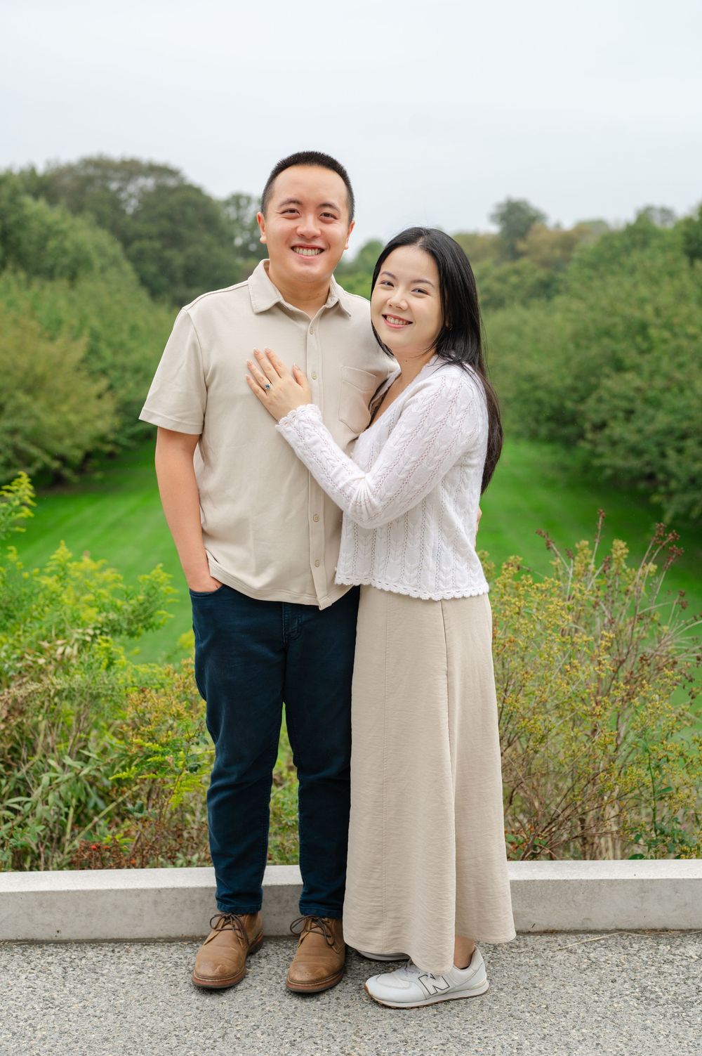 A smiling couple poses together in a lush green park, the woman in a white top and beige skirt resting her hand on the mans chest.