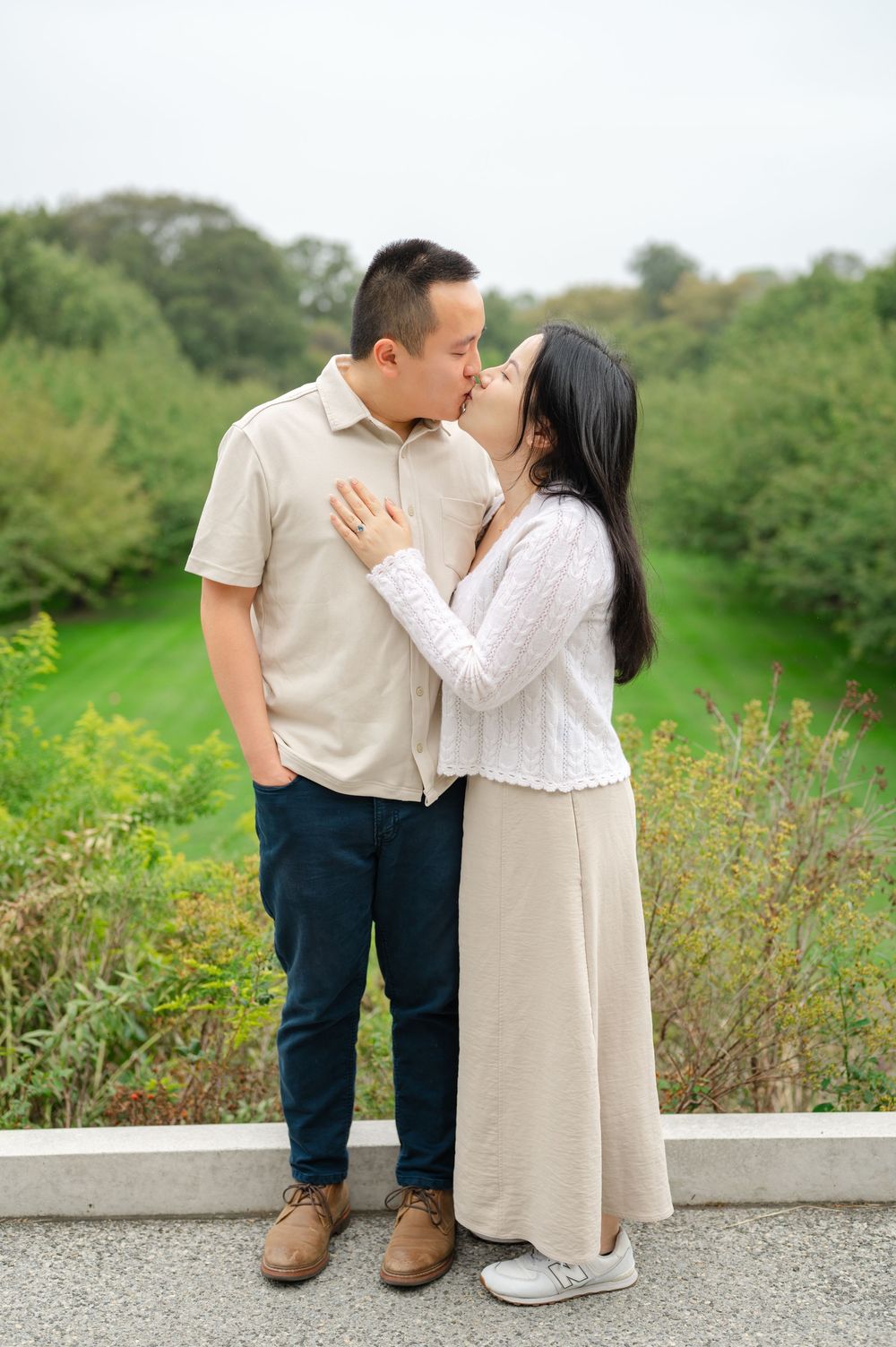 Asian couple sharing a tender kiss while standing on a path surrounded by greenery.